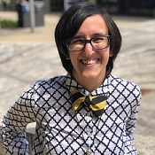 Nell, a white woman with short black hair, wears thick, black-rimmed glasses and smiles at the camera. She also wears a collared, button-up shirt that is white with a dark quatrefoil pattern; a black and gold diagonal stripe bow tie; and a glass pendant necklace. She is outside on a sunny day and leans back on the concrete structure behind her.